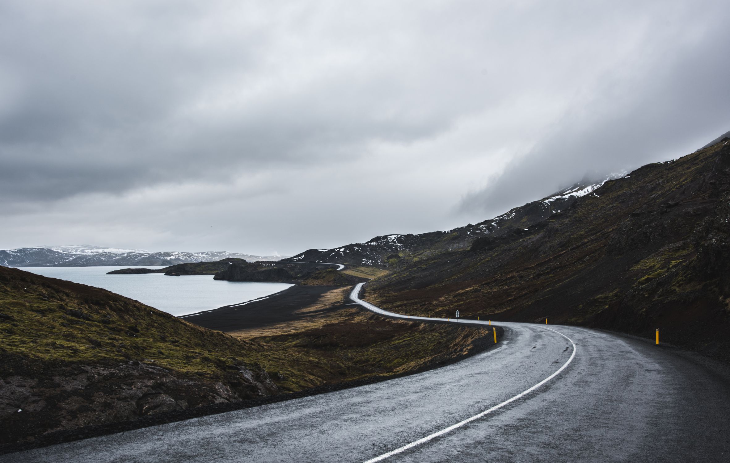 Winding road through Iceland's rugged landscape with cloudy skies and snowy mountains.