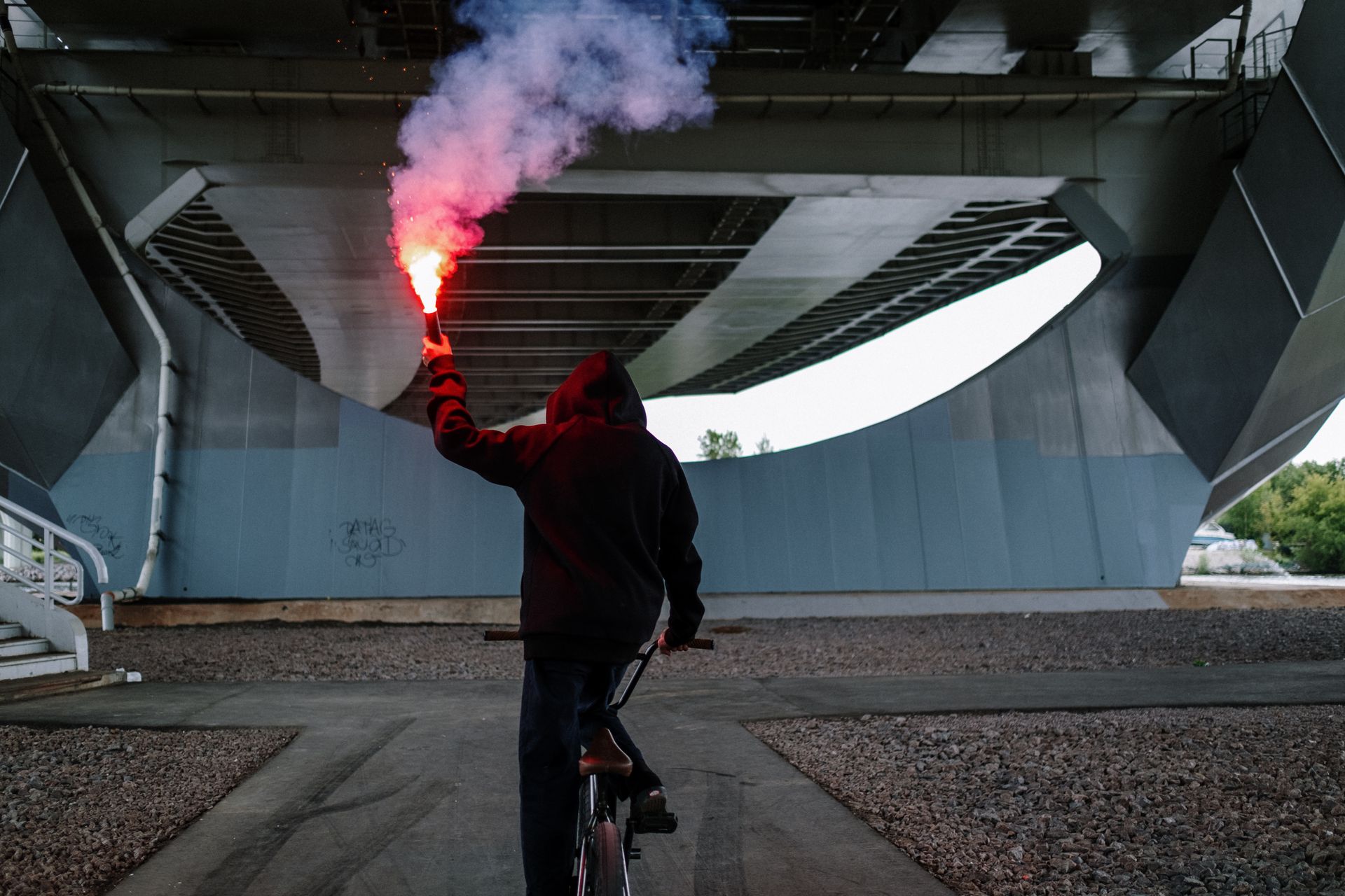 A man on a BMX bike holds a flare under an urban bridge, creating a dramatic urban scene.