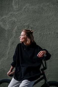 A woman in a black hoodie sits on a bicycle against a textured concrete wall, enjoying a moment of calm.