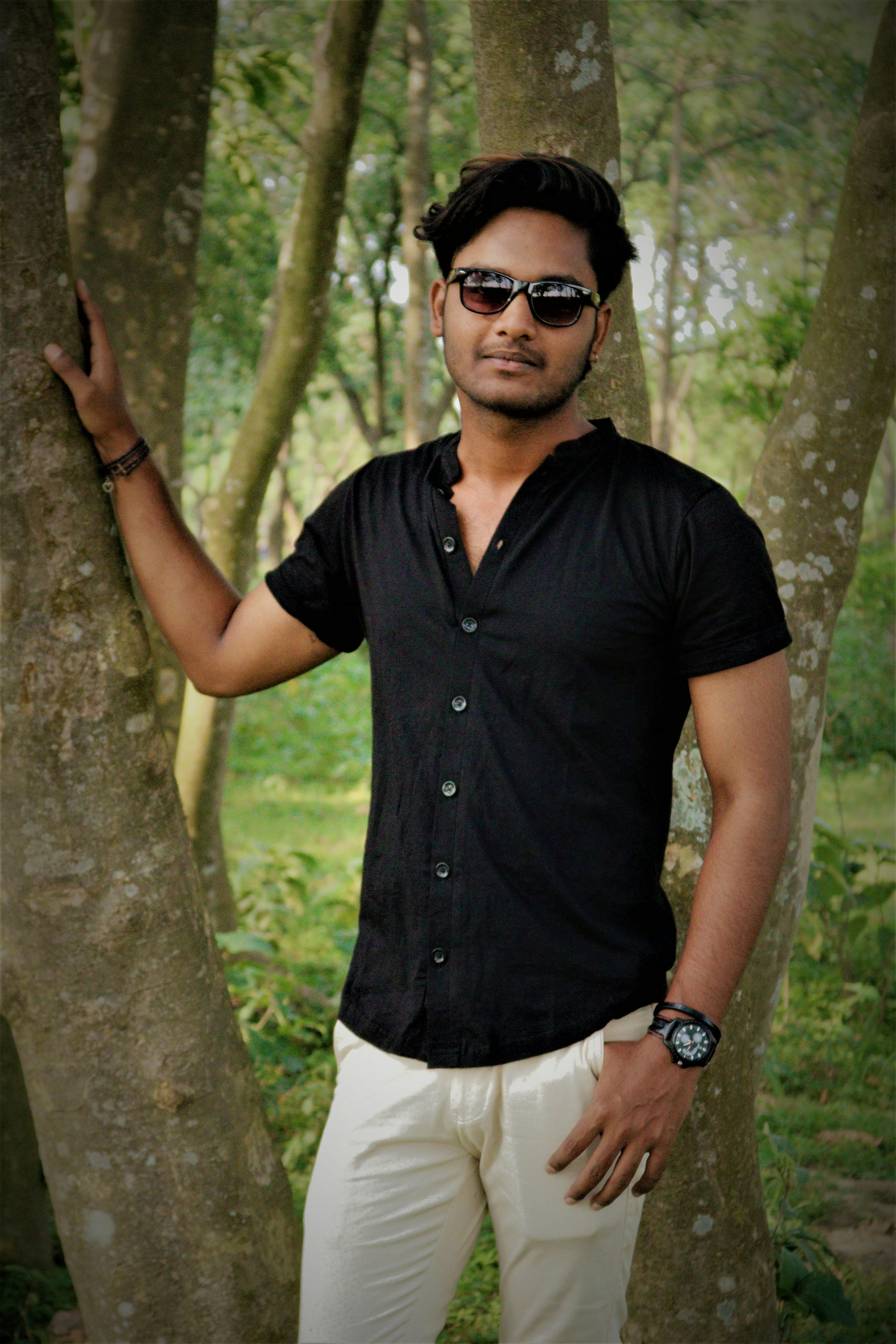Stylish man in black shirt posing in a lush park, exuding elegance and confidence.