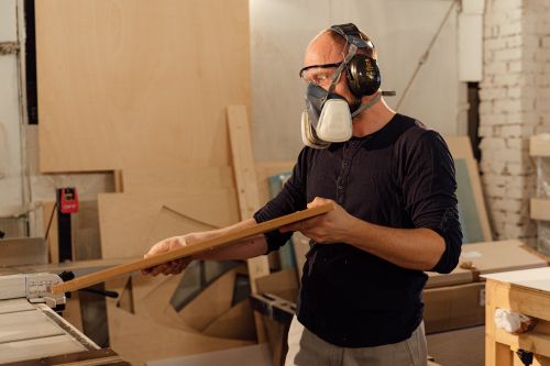 Free Carpenter wearing safety gear plans woodwork in a busy workshop environment. Stock Photo
