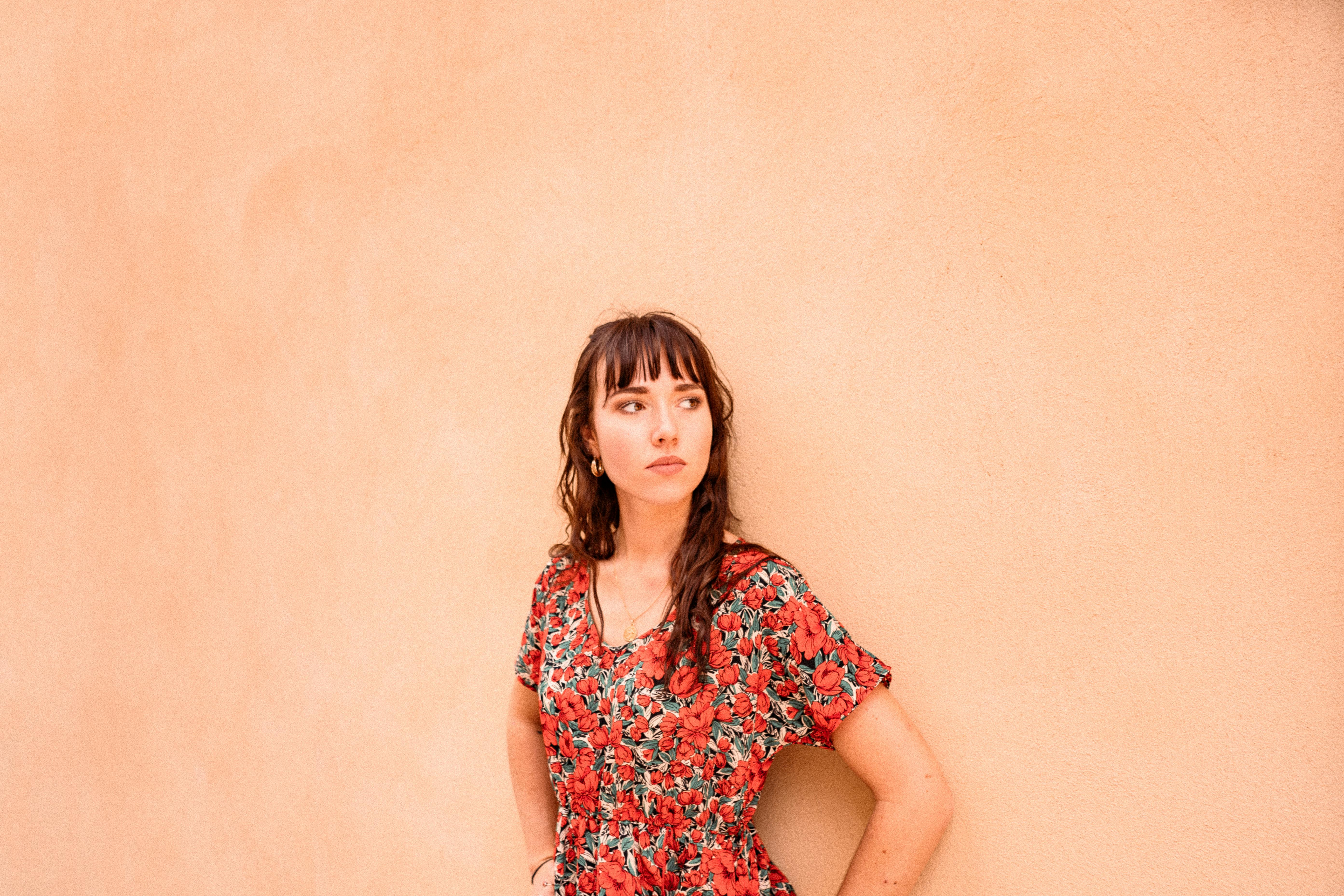 A young woman in a red floral dress leans against a peach wall, showcasing casual style.