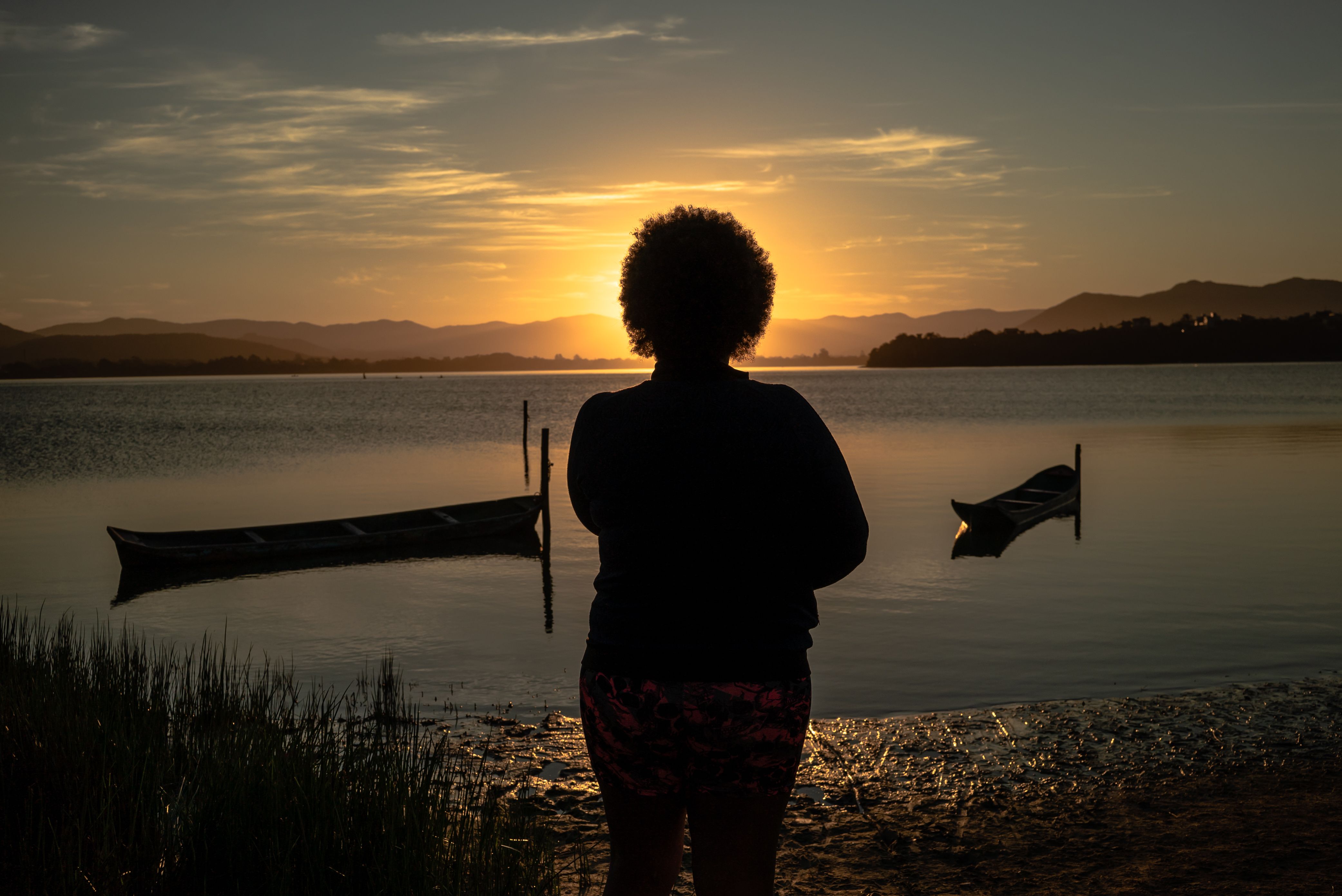 Silhouette of a woman standing by a lake, gazing at a tranquil sunrise with mountains in the background.