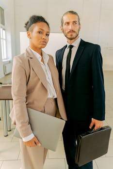 Two business professionals holding laptops, confident pose indoors.