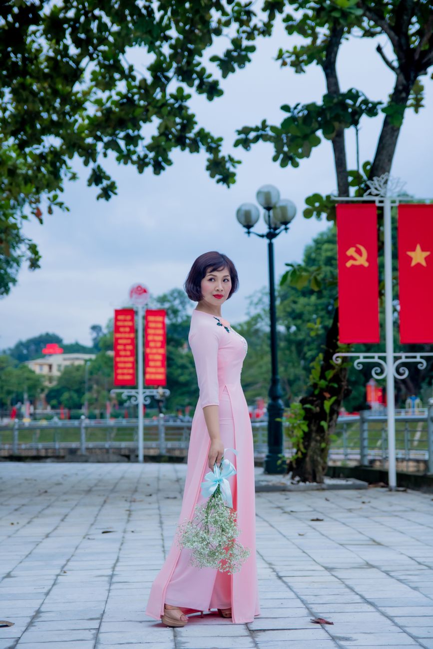 Asian woman in a pink dress poses with flowers on a tiled walkway surrounded by trees and red banners.