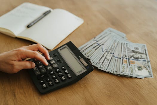 Close-up of a hand using a calculator with cash and a notebook on a wooden table.