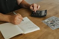 Person Writing on White Paper Beside Black Desk Calculator