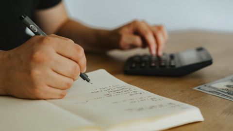 Close-up of hands writing calculations in a notebook with a calculator, focused on budgeting or financial work.