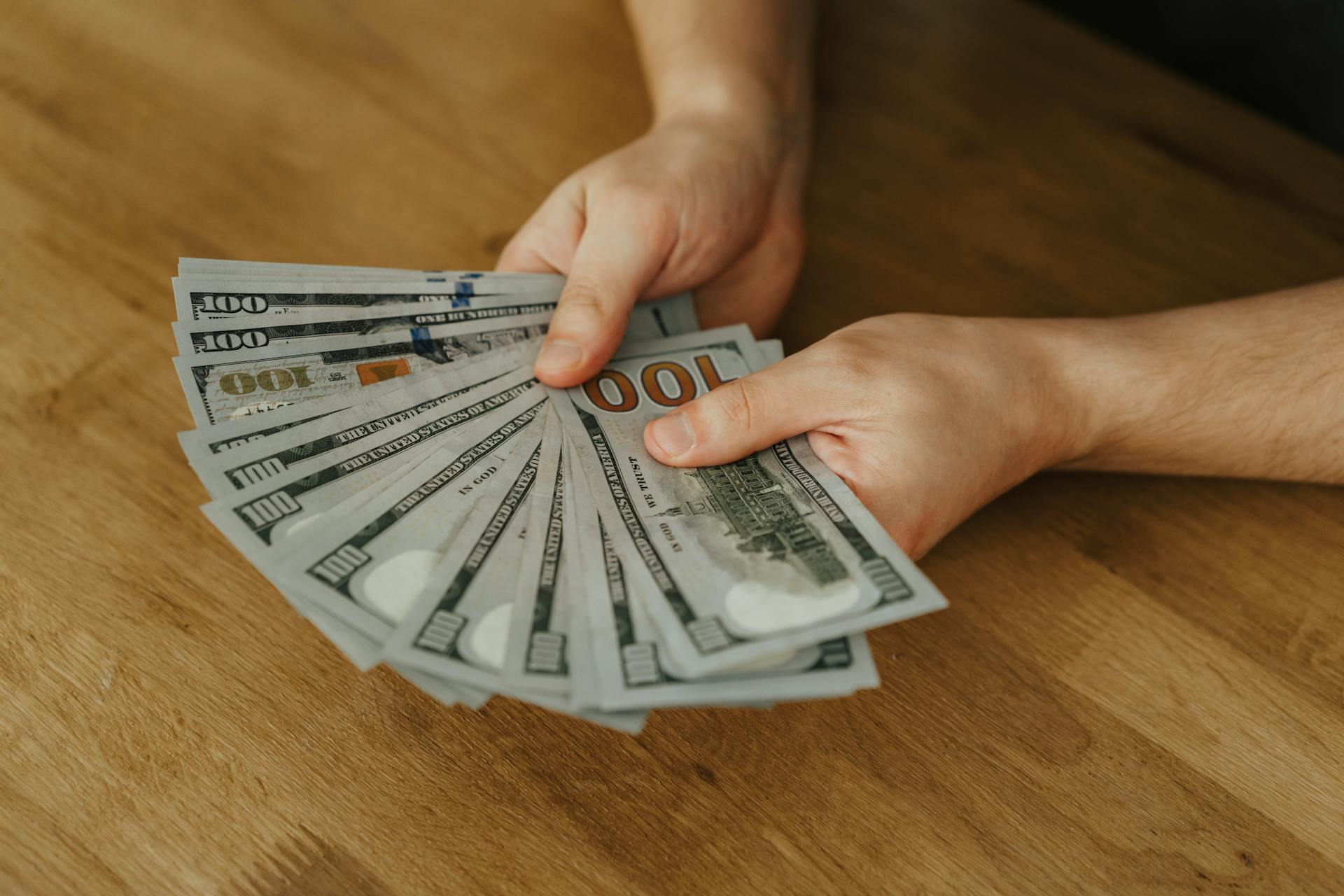 Close-up of hands holding several 100 US dollar bills on a wooden surface.