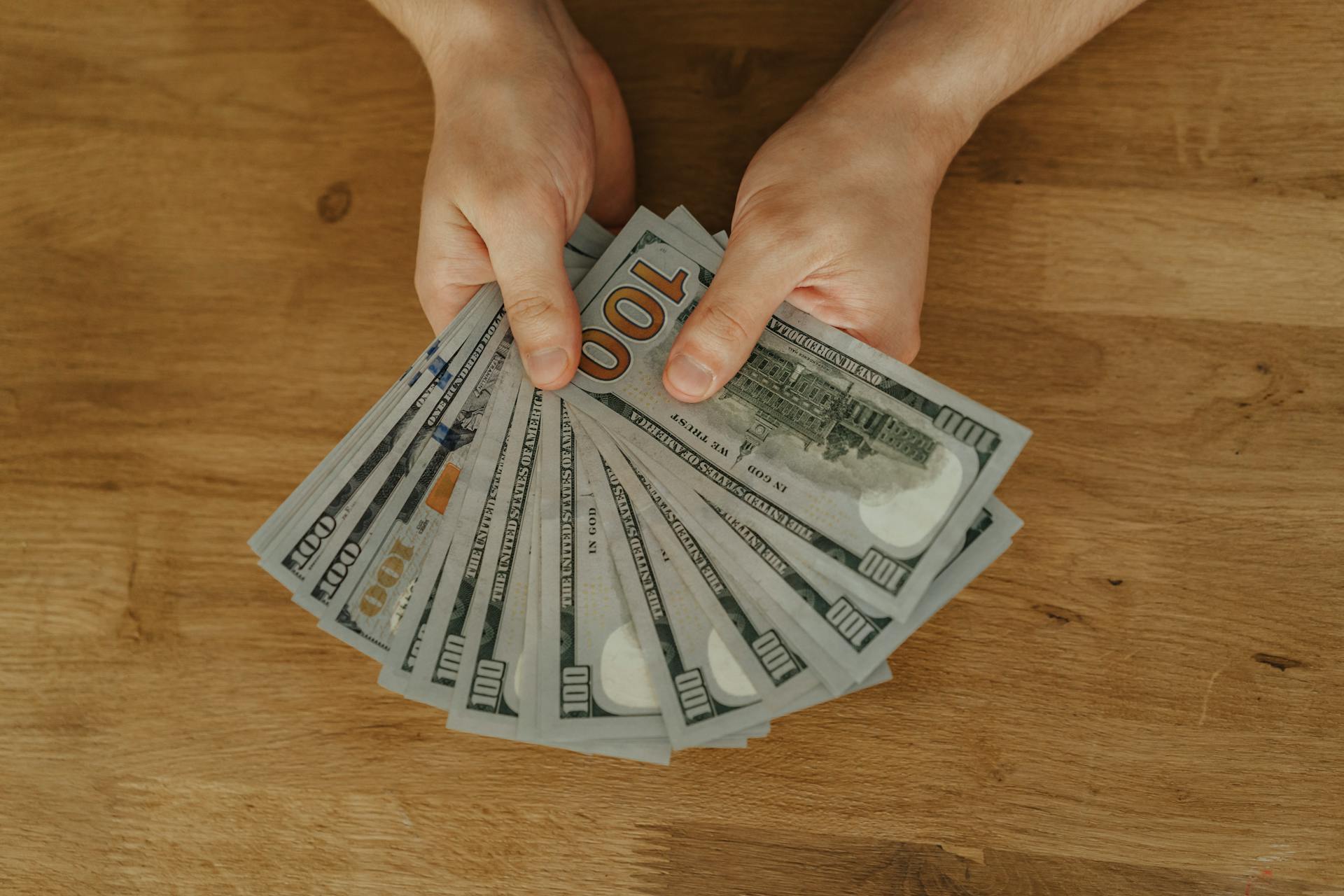 Close-up of hands fanning out US hundred dollar bills on a wooden table.