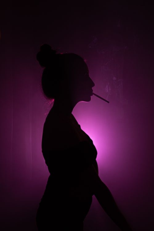 Free Silhouette of a woman smoking a cigarette against a purple backlight, creating a moody atmosphere. Stock Photo