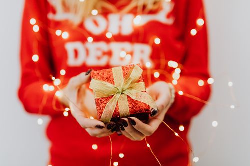Free Close-up of hands holding a gift box wrapped in red with festive lights and holiday sweater. Stock Photo
