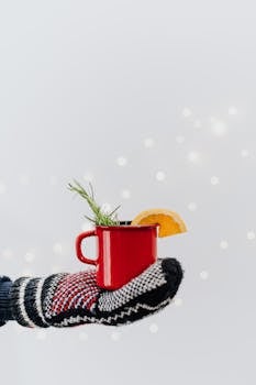 A festive red mug with an orange slice and rosemary held by a gloved hand in a winter setting.