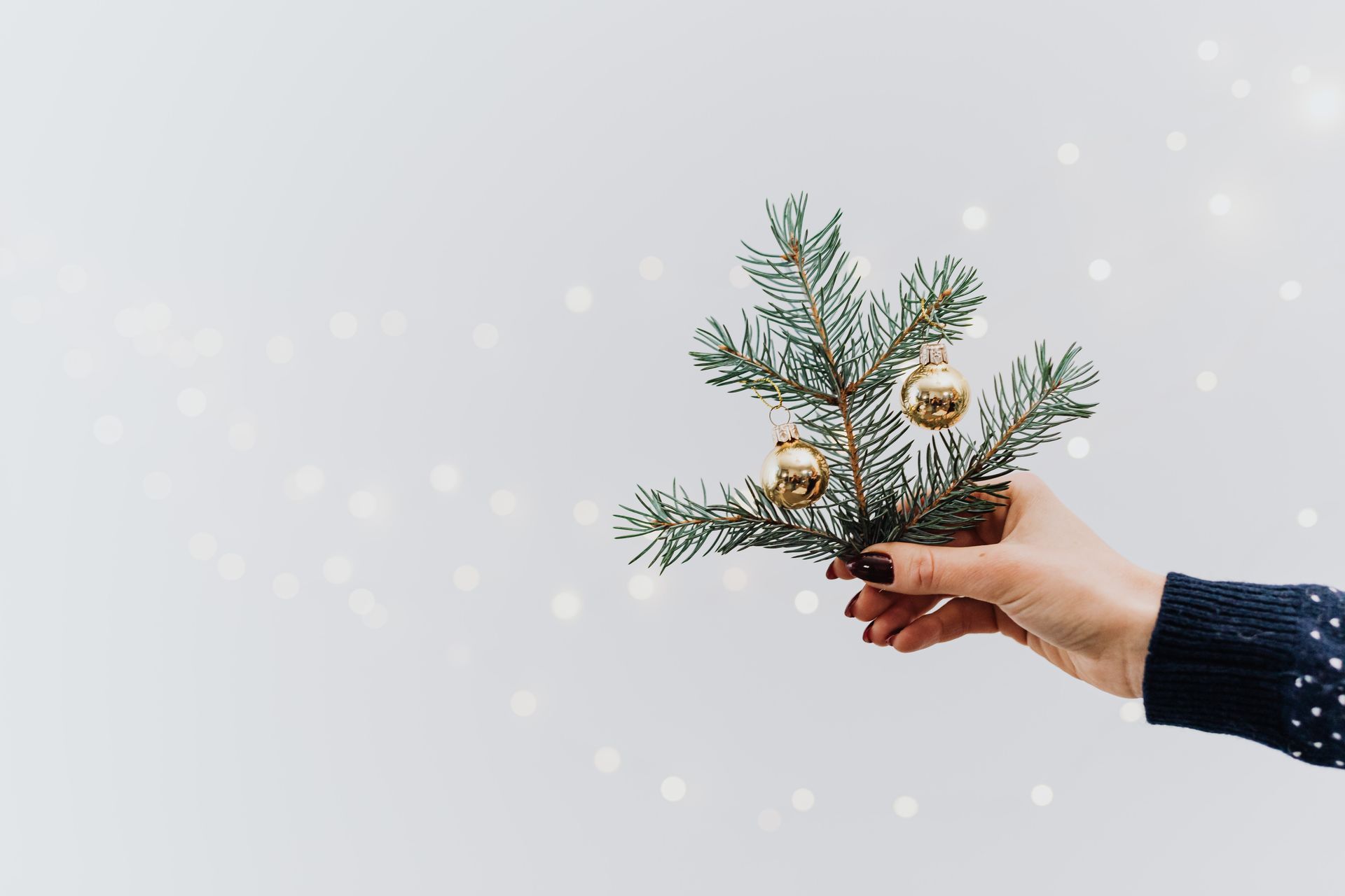 Elegant hand holding a pine twig with gold Christmas baubles and festive lights creating holiday spirit.