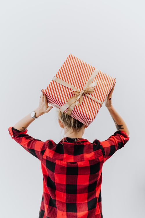 Free A woman in a plaid shirt holding a wrapped gift box on her head, conveying excitement and celebration. Stock Photo