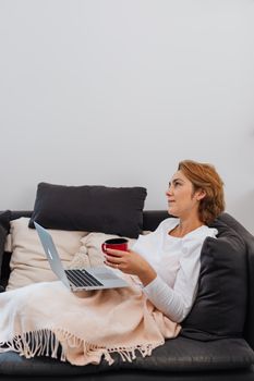 Woman enjoying leisure time with laptop and coffee on a cozy sofa indoors.