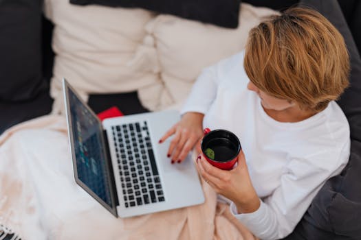 Woman in white top working on laptop and holding a mug, representing cozy remote work setup.