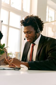 Formal attire man with headset working in bright office setting, writing notes.