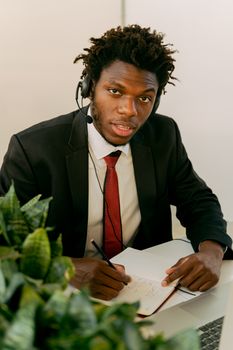 African American man wearing a headset, writing in a notebook at a workspace.
