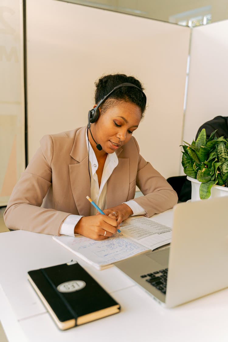 Woman In Beige Blazer Wearing Headset Writing On A Notebook 