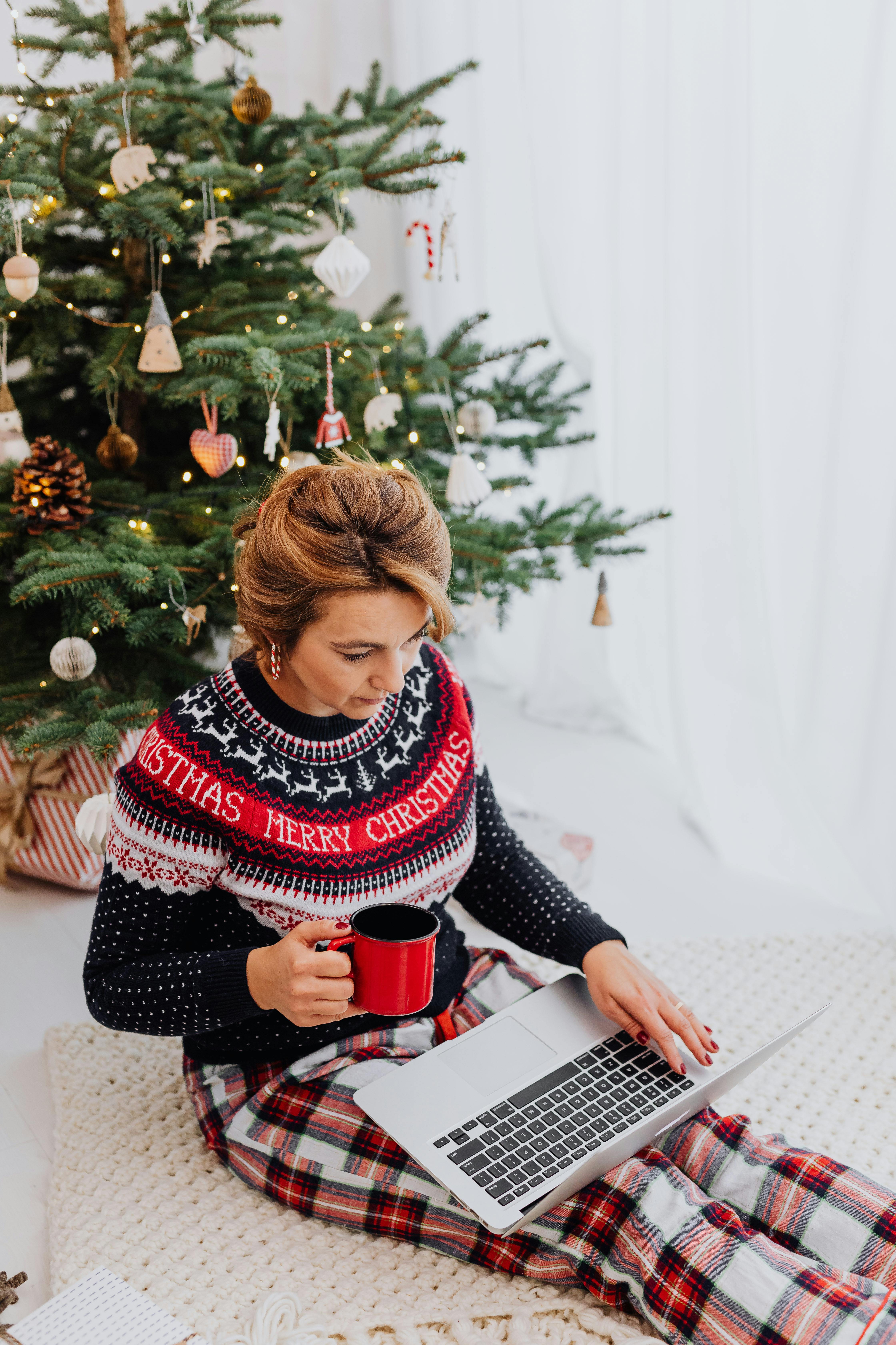在圣诞树旁，穿着毛衣的女人拿着杯子，正在使用笔记本电脑