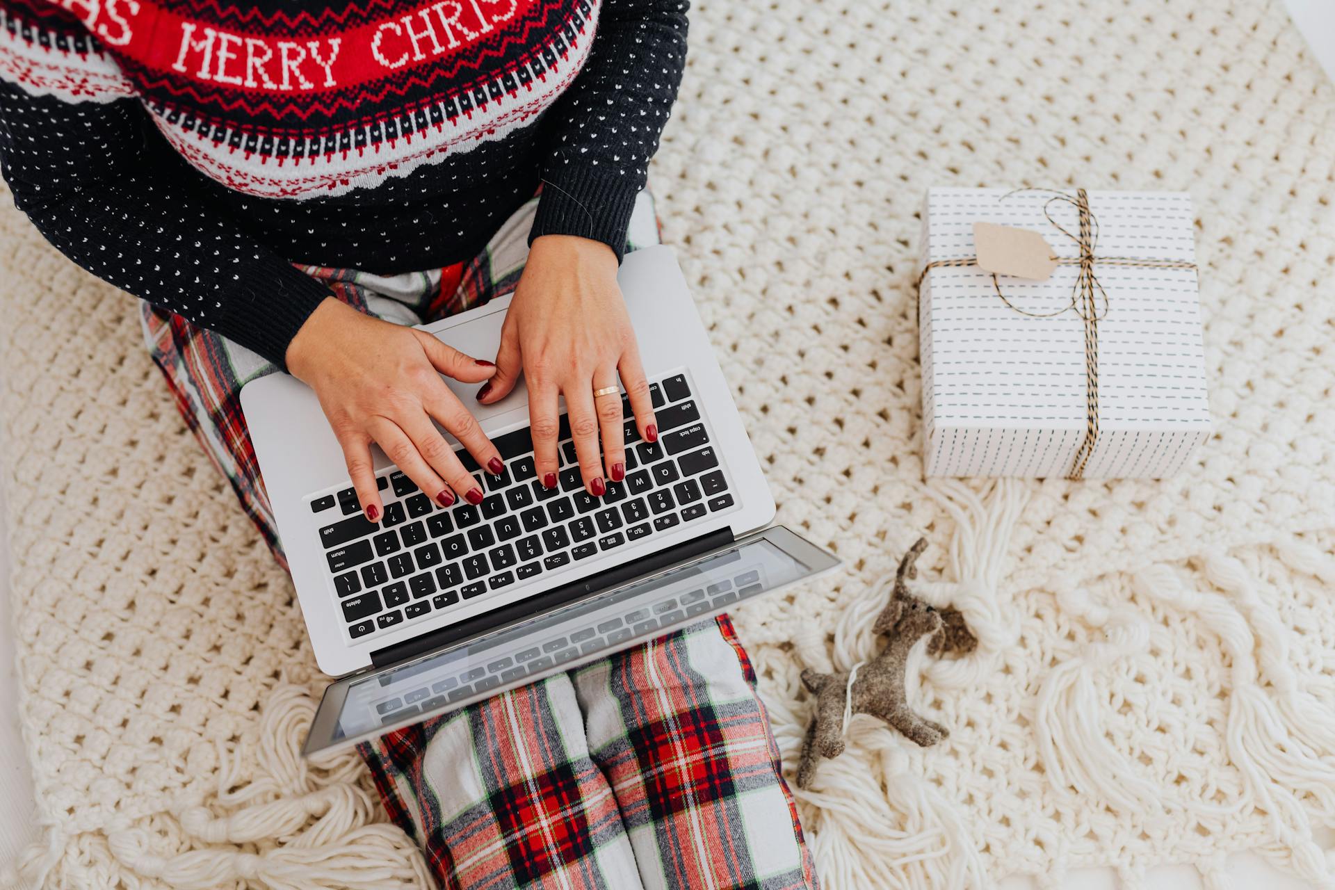 Woman working on a laptop with Christmas sweater and gift, capturing a cozy holiday work from home vibe.