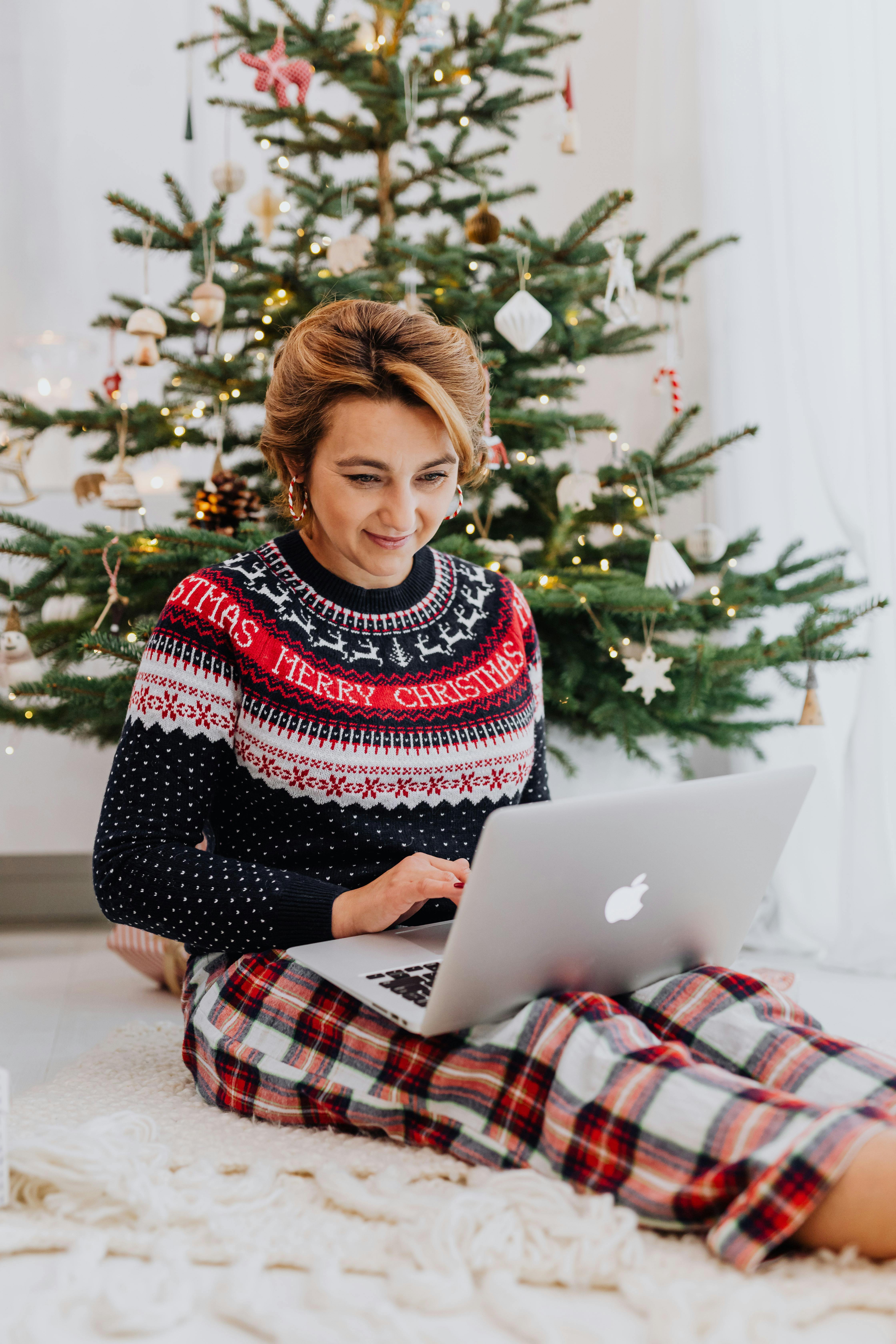 一名妇女穿着节日毛衣，在装饰好的圣诞树旁用笔记本电脑工作