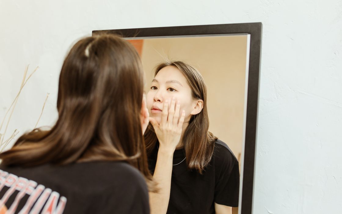 A woman studies her reflection in a mirror, touching her face.