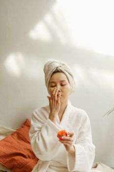 Asian woman in a robe applying skincare cream. Relaxing indoor beauty routine.
