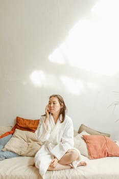 Asian woman in a bathrobe enjoying skincare routine in a sunlit room with cozy pillows.