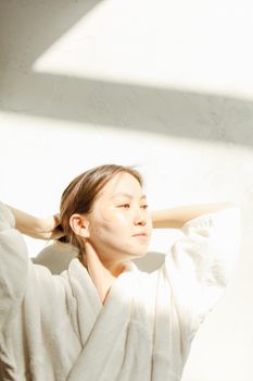 Asian woman enjoying morning skincare in soft sunlight, wearing a bathrobe.