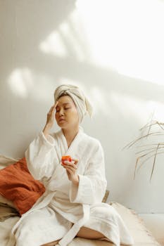Asian woman in robe applies skincare cream, promoting relaxation and beauty care.