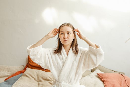 An Asian woman in a bathrobe applies skincare under eye masks at home.