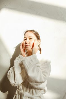 Woman in bathrobe applying skincare with natural light and shadows.