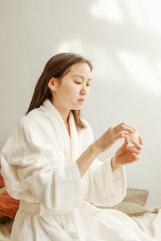 Asian woman in a white robe applying skincare cream, focusing on facial care indoors.