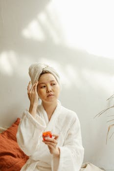Asian woman in bathrobe applying skincare cream indoors, enjoying self-care routine.