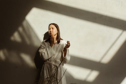 An Asian woman in a robe applies facial cream in soft, natural sunlight indoors.