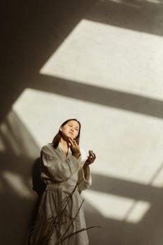 Asian woman applies skincare cream in sunlight with a serene expression indoors.