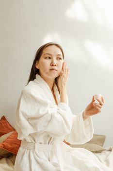 An Asian woman applies facial cream in a cozy, light-filled indoor setting.