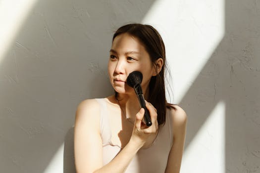 A young Asian woman applying makeup with a brush in natural lighting.