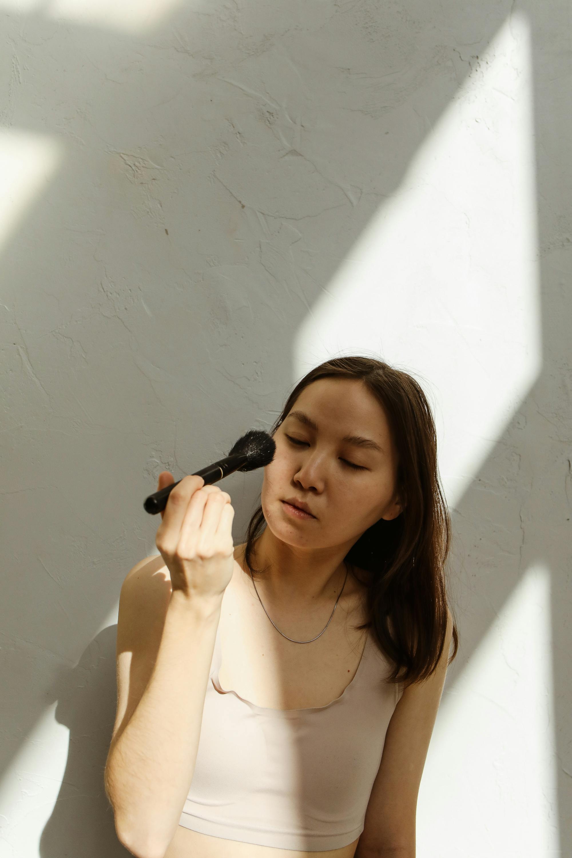 Young woman applying makeup in a sunlit room with soft shadows.