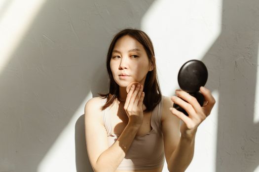 A young woman applying makeup, illuminated by natural light streaming through a window.
