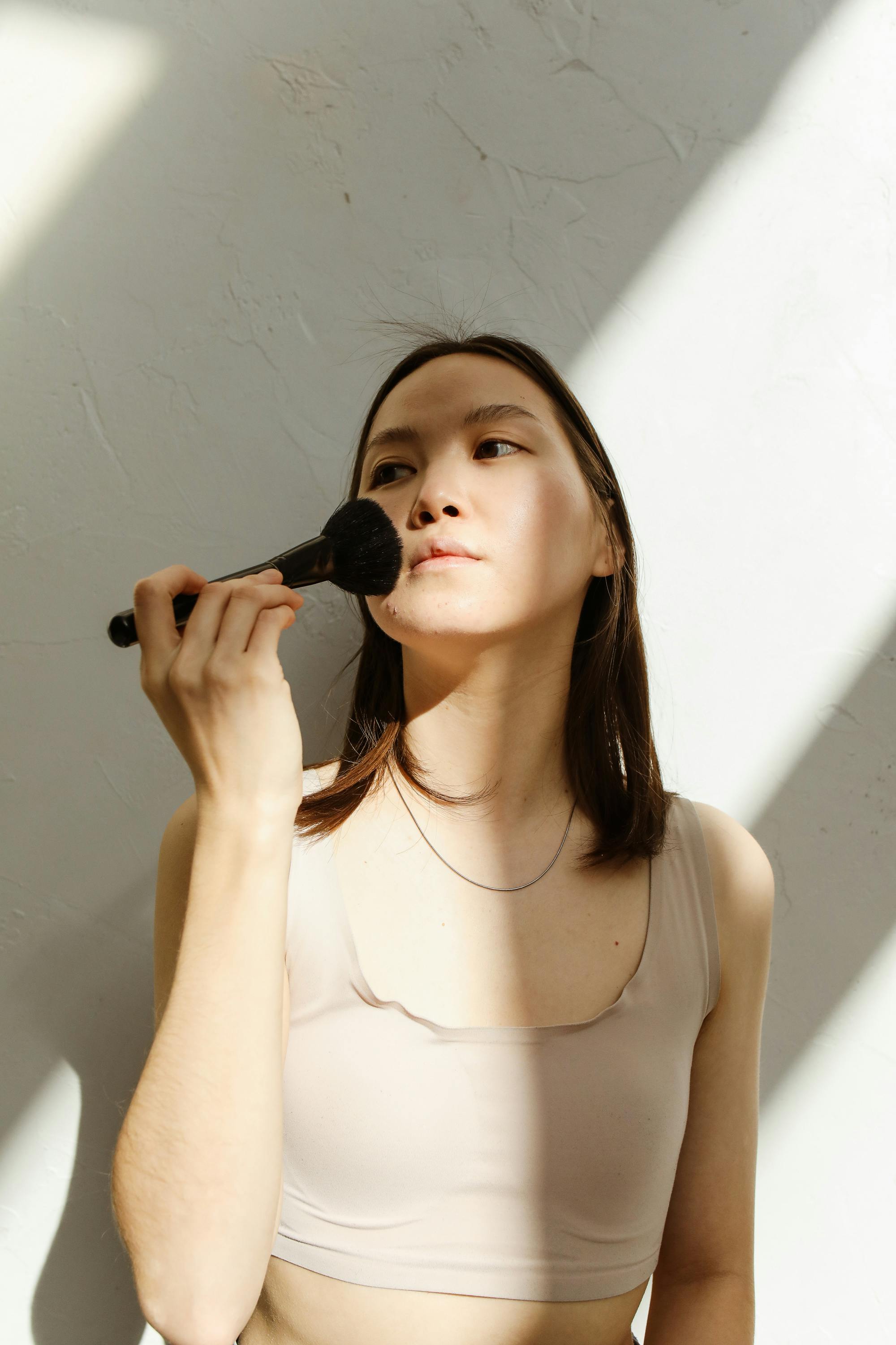 Portrait of a young woman using a makeup brush in soft natural light for beauty routine.