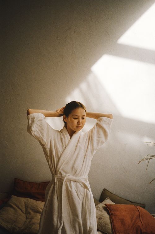 Free Asian woman in a bathrobe enjoying serene morning indoors, tying her hair. Stock Photo