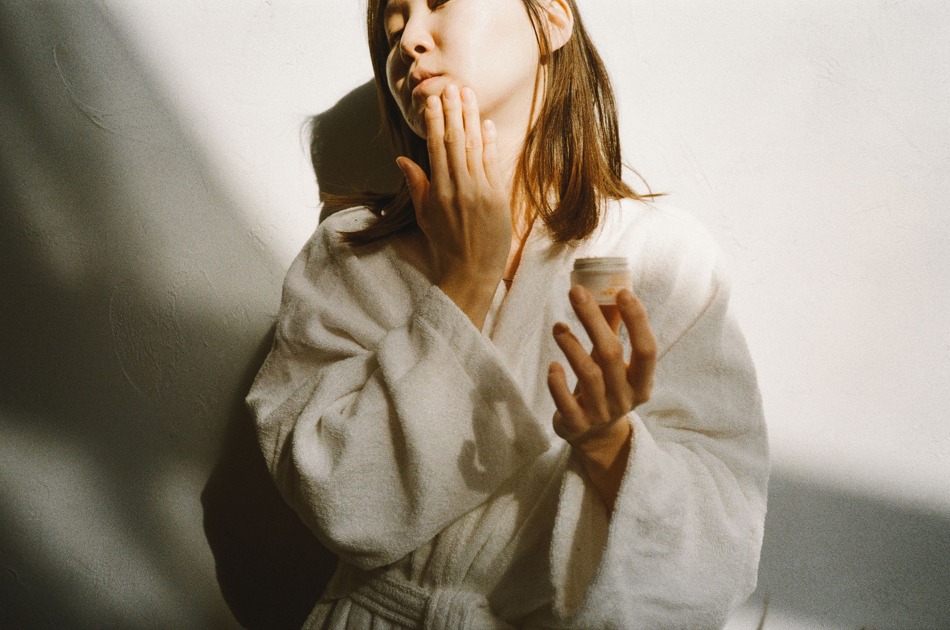 Asian woman in a white robe applying facial cream in a brightly lit indoor setting.