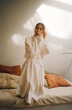 Asian woman in a bathrobe applying under eye masks for skincare in a cozy bedroom setting.