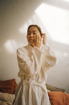 Asian woman in robe applies skincare under eye masks, bathed in gentle light.