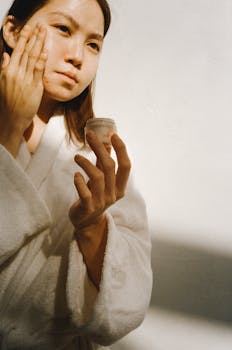 Young woman applying skincare moisturizer while wearing a bathrobe in natural light.