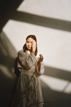 A young woman in a bathrobe applying moisturizer to her face in warm, soft lighting.