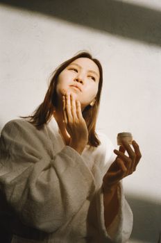 Asian woman in bathrobe applying moisturizer for her skincare routine. Bright sunlight illuminates the scene.